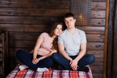 Young couple sitting crosslegs on plaid near dark wooden wall. Cosy countyside interior style, beautiful people in warm love relationsの写真素材