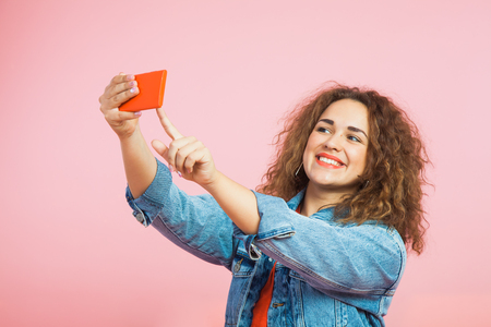 Beautiful girl with curly hair making selfie on smartphone. Woman in denim clothes photo heself on red mobile phone.の写真素材