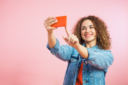 Beautiful girl with curly hair making selfie on smartphone. Woman in denim clothes photo heself on red mobile phone.の写真素材