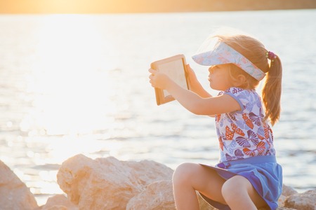 Cute little girl on rocky beach with digital tablet taking photos of a wonderful view. child is making selfie on a sunset or sunrise. Copy spaceの写真素材