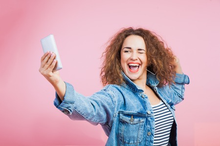 Beautiful expressive woman making selfie on smartphone. Female young model with curly hair at studio on pink background.の写真素材