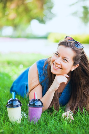 Young woman with sunglasses drink juicy tasty milkshakes on green lawn grass. Happy female model with two glasses of smoothie. Vertical shotの写真素材