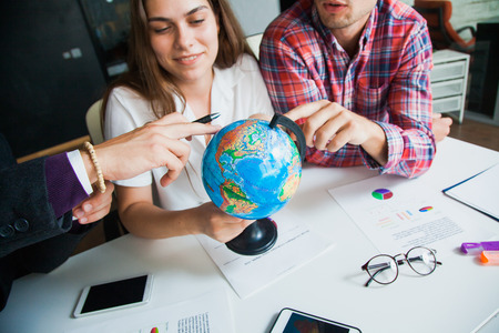 Group of three young people with globe choose country to travel to. Attractive woman squinted eyes to find a random place. Two guys support her. Travel worldwide conceptの写真素材