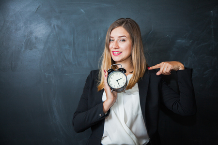 Business lady holding alarm clock with blackboard on background. Time management lack of free time concept. Pointing at watchesの写真素材