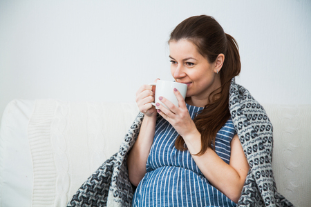 Pregnant woman drink tea and under warm cozy plaid at home on a winter evening.の写真素材