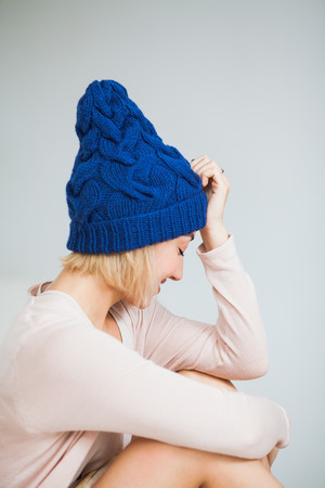 Young woman in warm dark blue hand knitted hat at home. Pretty lady in big blue cap, smiling happily. Vertical shot.の写真素材