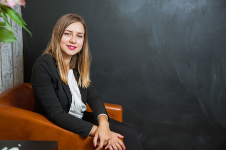 Young and beautiful businesswoman in armchair look at camera. Blackboard on background. Copy space for textの写真素材