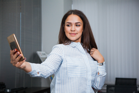 Attractive schoolgirl making selfie, female office worker using smart phone. Personal assistant at business center. Happy woman having video chatの写真素材
