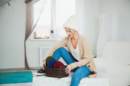 Beautiful woman in big white knitted hat indoors sit on chest with basket yarn. Knitting at home, hobby, cozy flat, denim casual clothing. Hygge, comfort and home concept,の写真素材
