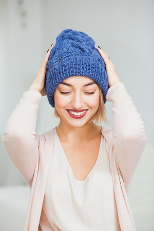 Young woman in warm dark blue hand knitted hat at home. Pretty lady in big blue cap, smiling happily. Vertical shot. Hygge, comfort and home concept.の写真素材