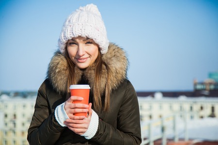 Cute young lady with coffe to go on a sunny frosty cold winter day. Woman in big cozy woolen hat holds hot drink warming frozen wrists.の写真素材