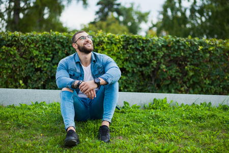 Young strong muscular guy sit on grass and relax. Male fitness model in jeans wear denim casual look, hipster man with beard and glasses.の写真素材