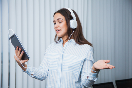 Pretty smiling businesswoman in white headphones holding digital tablet pc standing near big office window, having video conference, using business apps, internet banking, browsing web serviceの写真素材