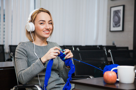 Young woman knitting at working place wearing headphones and smile. Leisure time at office concept. Businesswoman procrastinateの写真素材