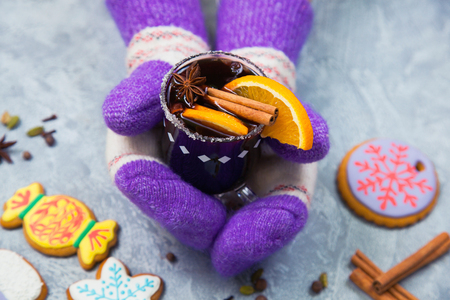 Mulled wine in glass mug with spices, female hands in knitted mittens. Christmas hot drink with decorated new year ginger bread on gray stone table. Top view.の写真素材