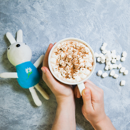 female hands hold big mug with cacao and marshmallow on gray background. Child toy, copy spaceの写真素材