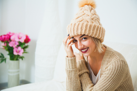 Hygge, comfort and home concept. Young woman in warm beige hand knitted hat at home. Pretty lady in big wool cap, smiling happily. Turn away from camera. Copy spaceの写真素材