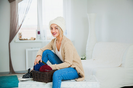 Beautiful woman in big white knitted hat indoors sit on chest with basket yarn. Knitting at home, hobby, cozy flat, denim casual clothing.の写真素材