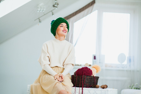 Young woman in warm hand knitted hat at home. Pretty lady in big green cap, smiling happily. Beautiful girl knitting in cozy flat wearing casual clothes. Horizontall shot. Copy spaceの写真素材