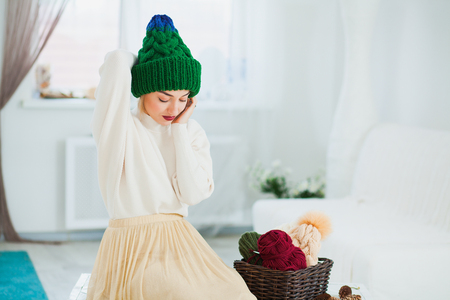 Young woman in warm hand knitted hat at home. Pretty lady in big green cap, smiling happily. Beautiful girl knitting in cozy flat wearing casual clothes. Horizontall shot. Copy spaceの写真素材