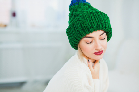 Young woman in warm hand knitted hat at home. Pretty lady in big green cap, smiling happily. Horizontall shot. Copy spaceの写真素材