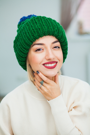 Young woman in warm hand knitted hat at home. Pretty lady in big green cap, smiling happily. Vertical shot. Hygge, comfort and home concept.の写真素材