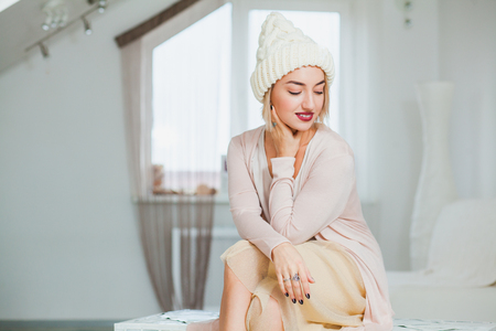 Young woman in warm hand knitted hat at home. Beautiful lady in big white knitted hat indoors sit on chest with basket yarn. Knitting at home, hobby, cozy flat, denim casual clothing.の写真素材