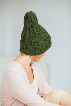 Young woman in warm hand knitted hat at home. Pretty lady in big green cap, smiling happily. Vertical shot. Hygge, comfort and home concept.の写真素材