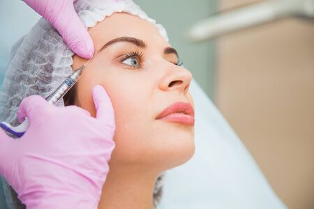 Young woman at cosmetologist office remove wrinkles on face contour. Toxin injections, anti-age procedure, skincare concept. Doctor and patient.の写真素材