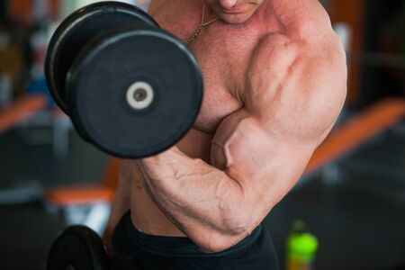 Bodybuilder at well-equipped gym lifting heavy dumbbells. Muscular athlete exercise on biceps. Healthy, active lifestyle concept. Closeup of sportsman arm.の写真素材