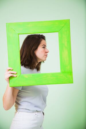 Young woman with green wooden frame on light background. Vertical portrait of casual female person, no retoching.の写真素材