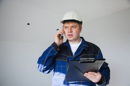 Builder foreman in a white helmet at a construction site indoors, architect flat renovation, professional foreman at work discussing by smartphoneの写真素材