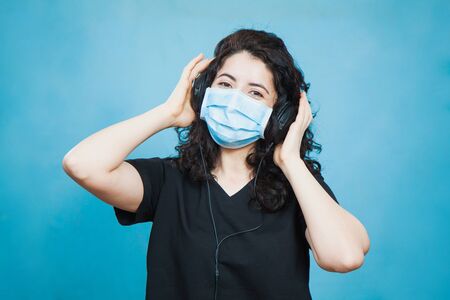 Cheerful culry haired woman in antiviral mask wears headphones and listen to music at home. Social distance, quarantine isolation, virus prevention concept.の写真素材