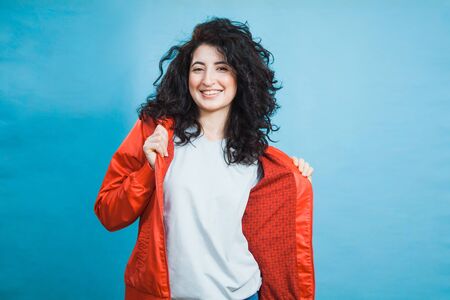 Happy pretty young girl in red coat with dark curly hair and open smile. Portrait of smiling woman on blue background with copy space for some text.の写真素材