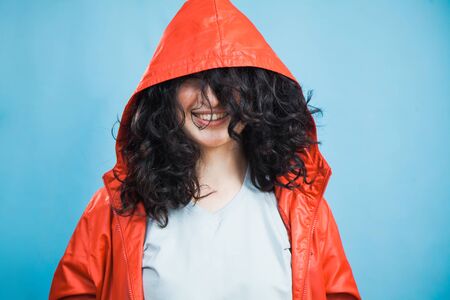 Happy pretty young girl in red coat hood with dark curly hair and open smile. Portrait of smiling woman with hood on her face on blue background with copy space for some text.の写真素材