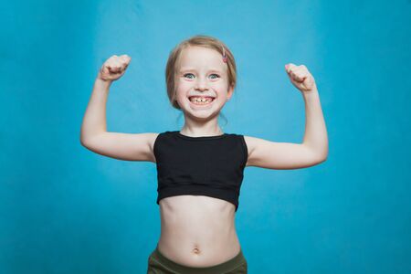 Happy pretten girl do sports and show muscles biceps on blue background. Confident child dream to be fit, slim and healthy. Fitness, gymnastics and active lifestyle concept.の写真素材