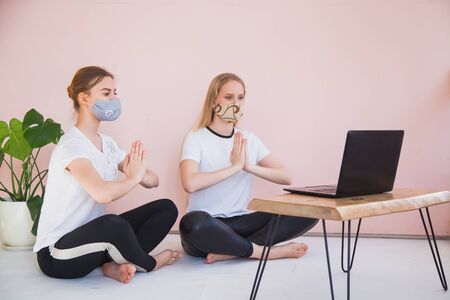 Two sisters doing yoga at home with laptop on quarantine. Online translation by internet, girls in protective masks, hands namaste, remote training on covid-19 outbreak. Staying at home concept.の写真素材