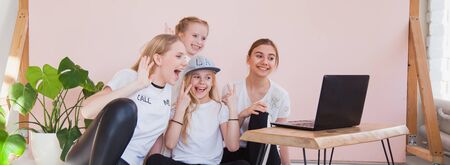 Four young sisters smile and look at monitor of laptop. Family use internet chat on quarantine, social life online, home isolation concept. Wide bannerの写真素材