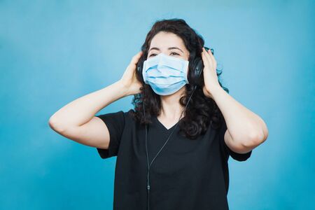 Cheerful culry haired woman in antiviral mask wears headphones and listen to music at home. Social distance, quarantine isolation, virus prevention concept.の写真素材