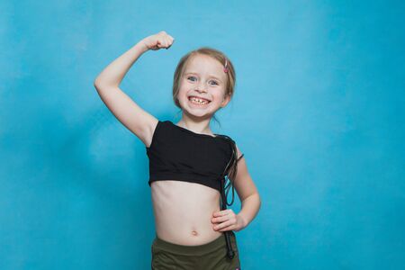 Happy pretten girl do sports and show muscles biceps on blue background. Confident child dream to be fit, slim and healthy. Fitness, gymnastics and active lifestyle concept.