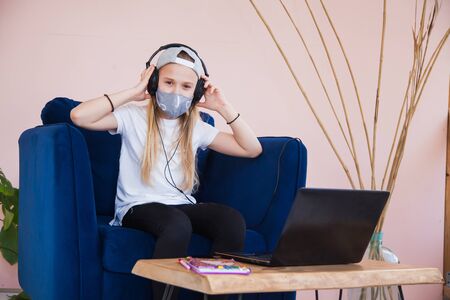 Teenage girl study at home with headphones and laptop. Young female student homeschooling during quarantine. Protective antiviral mask, social distance, self-isolation concept.の写真素材