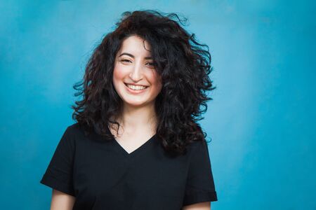 Portrait of a happy armenian woman with dark curly hair. Beautiful young female model in casual t-shirt smiling on blue background. Copy space for text.の写真素材