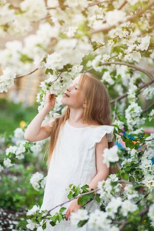 Happy little blonde girl in blooming apple tree. Female child in white dress smell apple flower blossoms in garden.の写真素材
