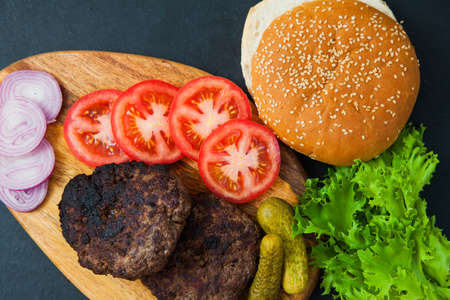 Set of beef burger ingredients on dark table. two cutlets, tomatoes, onions, salad and fresh sesame buns. Delicious homemade dish.の写真素材
