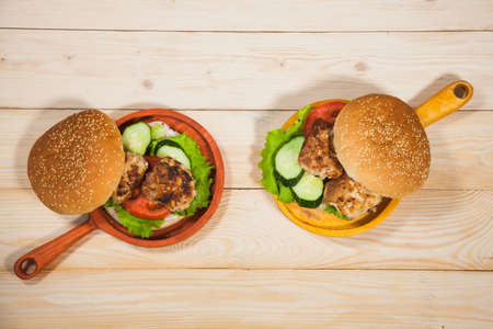 Two homemade hamburgers with fresh vegetables and chicken cutlets. Pair of burgers on handmade wooden serving pans. Delicious food on wooden table.の写真素材