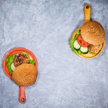 Two homemade hamburgers with fresh vegetables and chicken cutlets. Pair of burgers on handmade wooden serving pans. Delicious food on gray table.の写真素材
