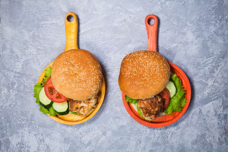 Two homemade hamburgers with fresh vegetables and chicken cutlets. Pair of burgers on handmade wooden serving pans. Delicious food on gray table.の写真素材