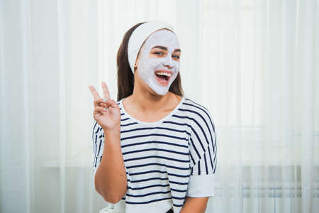 Happy and beautiful girl with white clay mask on her face. Home beauty treatment. Skincare and rejuvenation concept.の写真素材