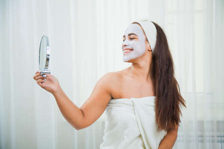 Happy woman in towel and facial mask look in the mirror. Home beauty treatment. Skincare and rejuvenation concept.の写真素材