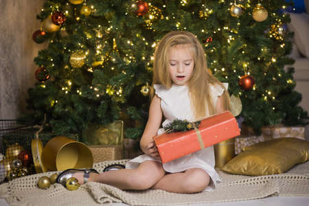 Happy cute girl sit near christmas tree with red gift box. New year holiday, female child with present in festive decorated home room.の写真素材